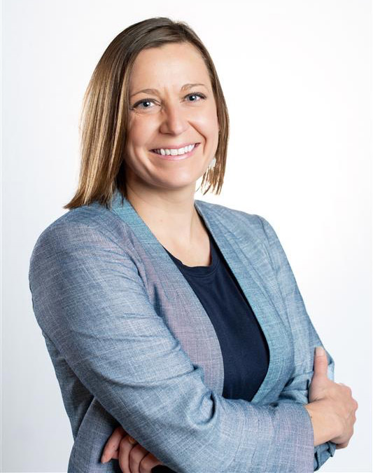 Head shot of Erika Debrick Kelly. She is a blonde woman and is wearing a dark blue shirt with a light blue blazer. She is smiling.
