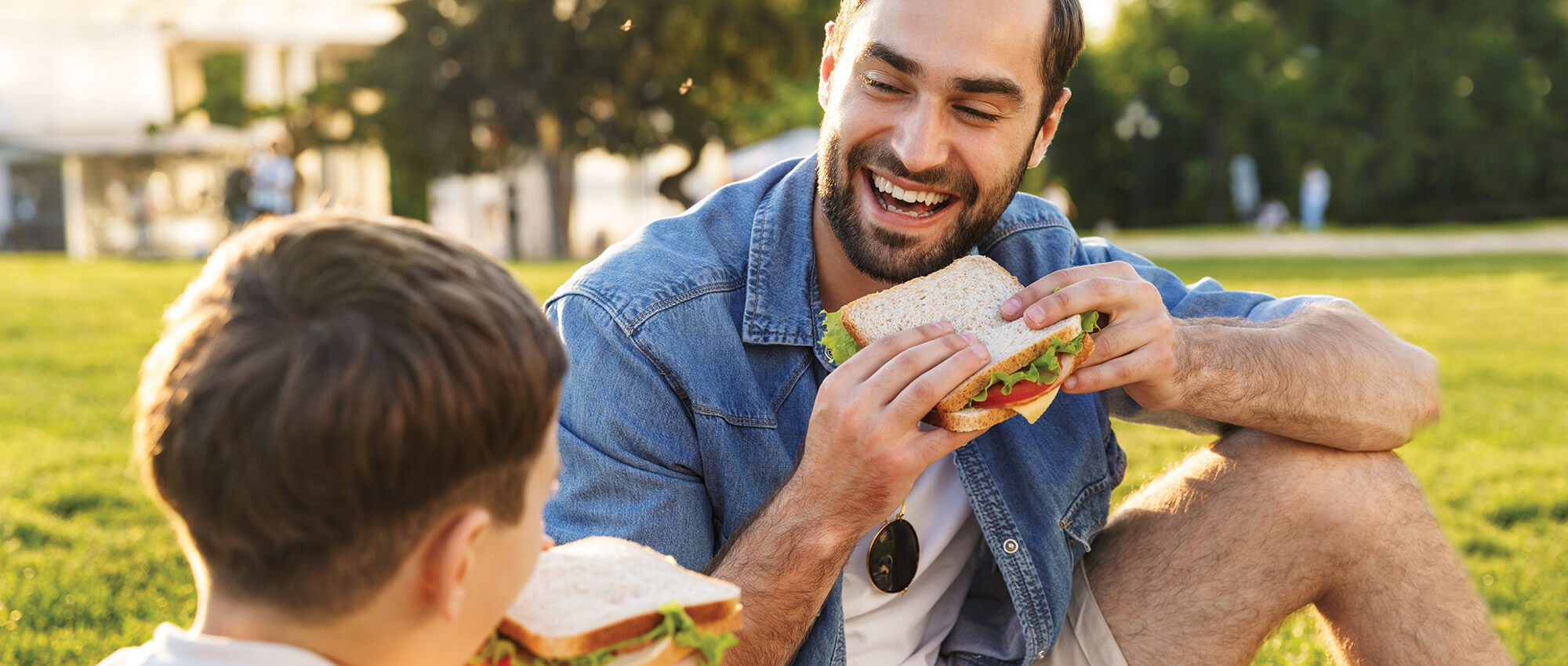 A man and his son are sitting in the park, smiling and eating sandwiches.